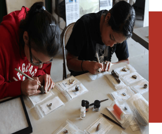 Two women labeling artifacts using a micro pen. Artifacts are also on a table where they are sitting. Arifacts are in archival bags. 